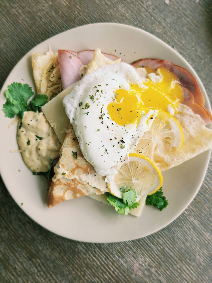 The frenchie on a pale plate — a folded savory crêpe with ham and gruyère, a nested fried egg with a runny yolk on top, dijon dressing, lemon slices and fresh cilantro