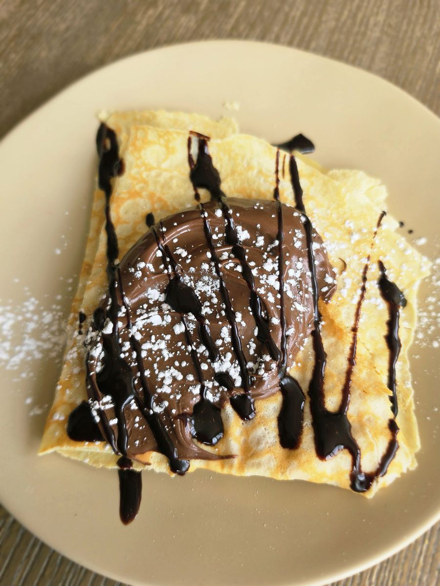 The kids just nutella crêpe on a pale plate — a folded crêpe spread with nutella, finished with chocolate drizzle and powdered sugar