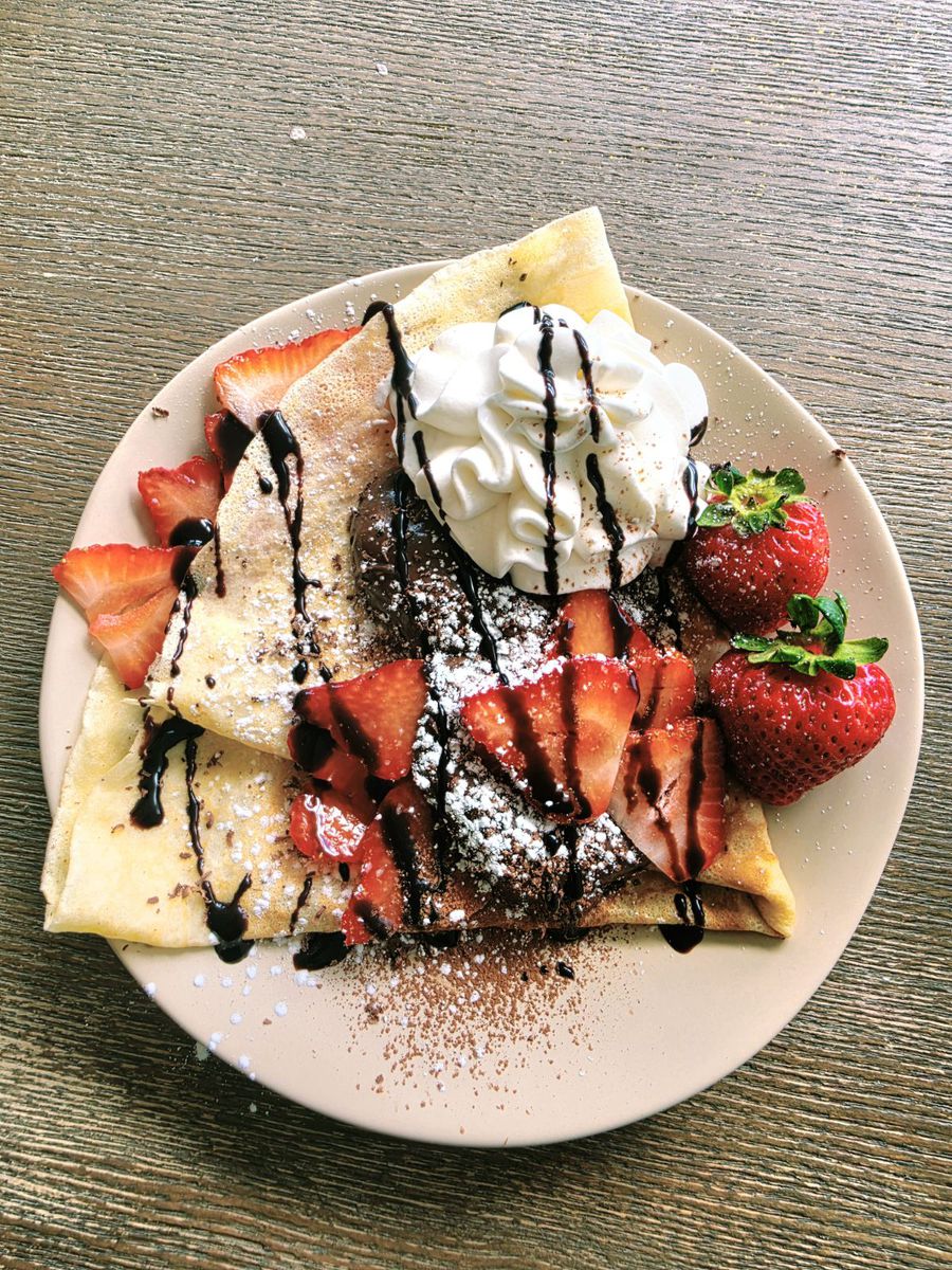 The nutella + fruit crêpe on a pale plate — folded sweet crêpe with nutella and sliced strawberries, topped with a swirl of chantilly cream, chocolate syrup and powdered sugar