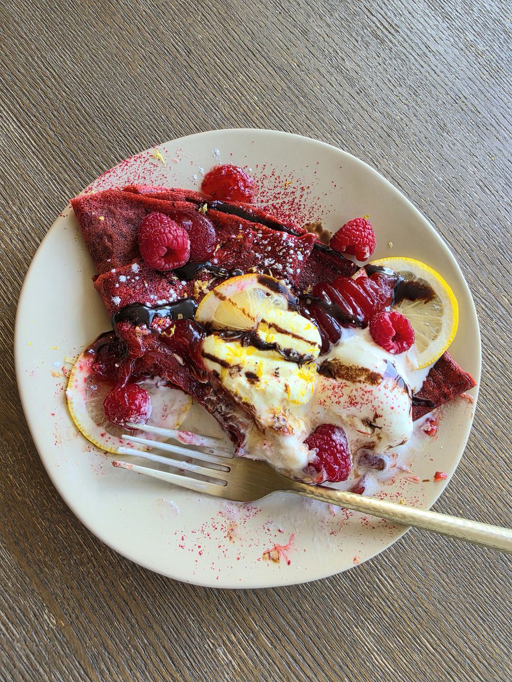 The poppy crêpe on a pale plate — a folded red velvet poppyseed crêpe layered with lemon cheesecake cream and raspberry compote, topped with fresh raspberries, candied lemon slices, dark chocolate drizzle and raspberry powder