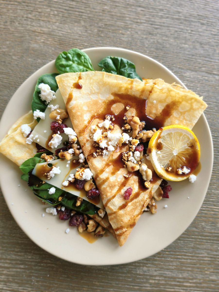 The veggie crêpe on a pale plate — a folded crêpe over swiss cheese and fresh spinach, scattered with crumbled feta, walnuts and dried cranberries, finished with balsamic glaze and a lemon slice