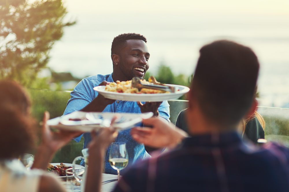 A man at a sunlit outdoor table passing a large platter of food across to other guests