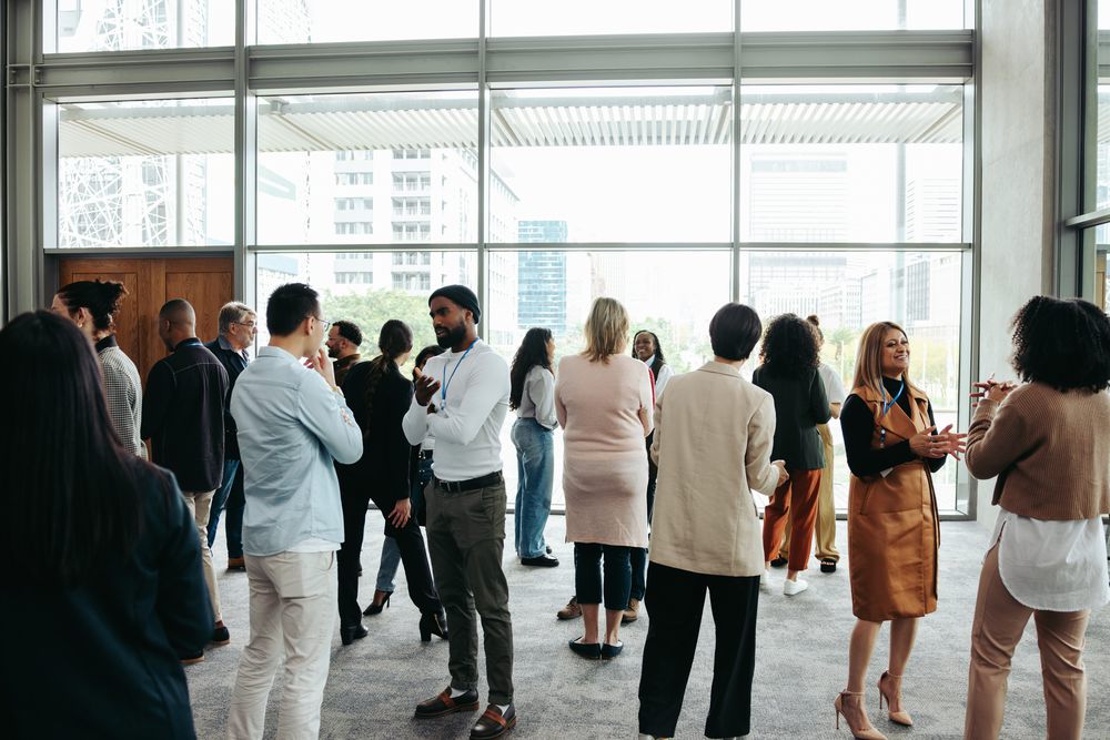 Colleagues standing and talking in small groups at a bright office event space with floor-to-ceiling windows