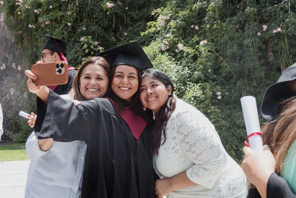 A graduate in cap and gown leaning in for a selfie with two family members outdoors after the ceremony