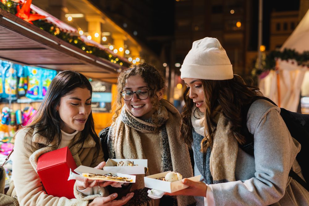 Three friends in coats and scarves at a lit-up winter market, holding open boxes of crêpes topped with ice cream