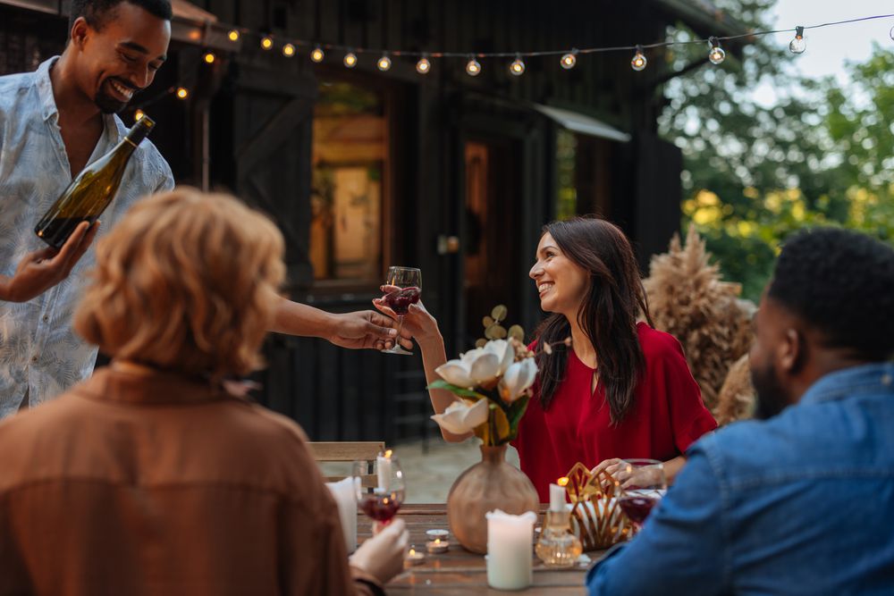 Friends around an outdoor table at dusk under string lights, passing a glass of wine