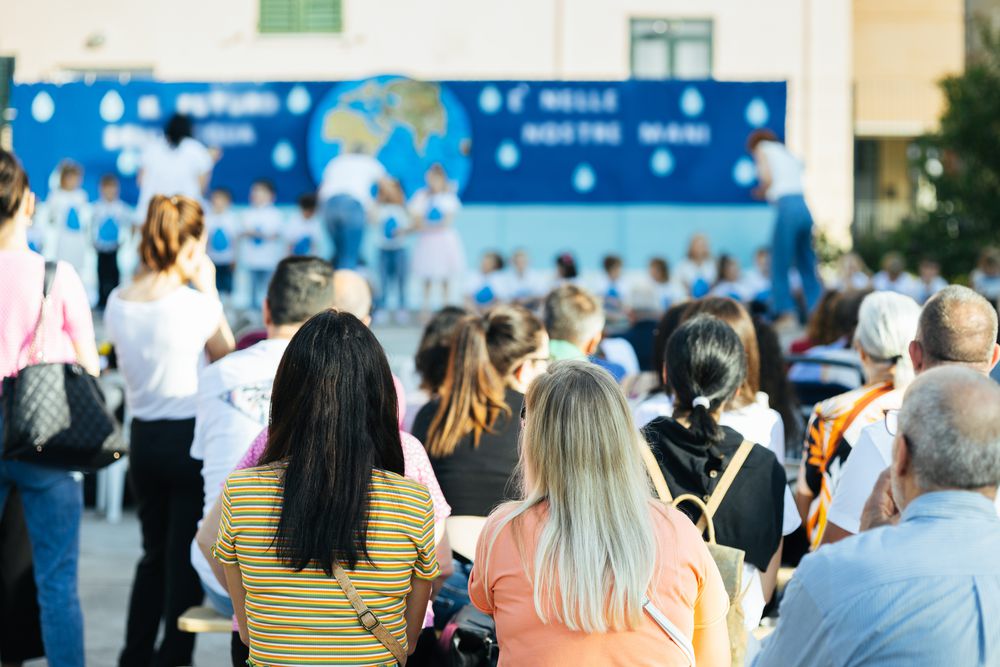 Parents and families seated outdoors watching children perform on a stage at a school event