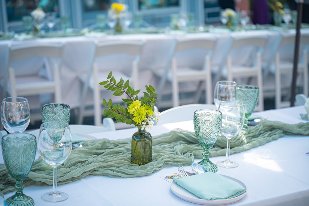 A long white reception table set with sage runners, patterned green glassware and a small floral centrepiece