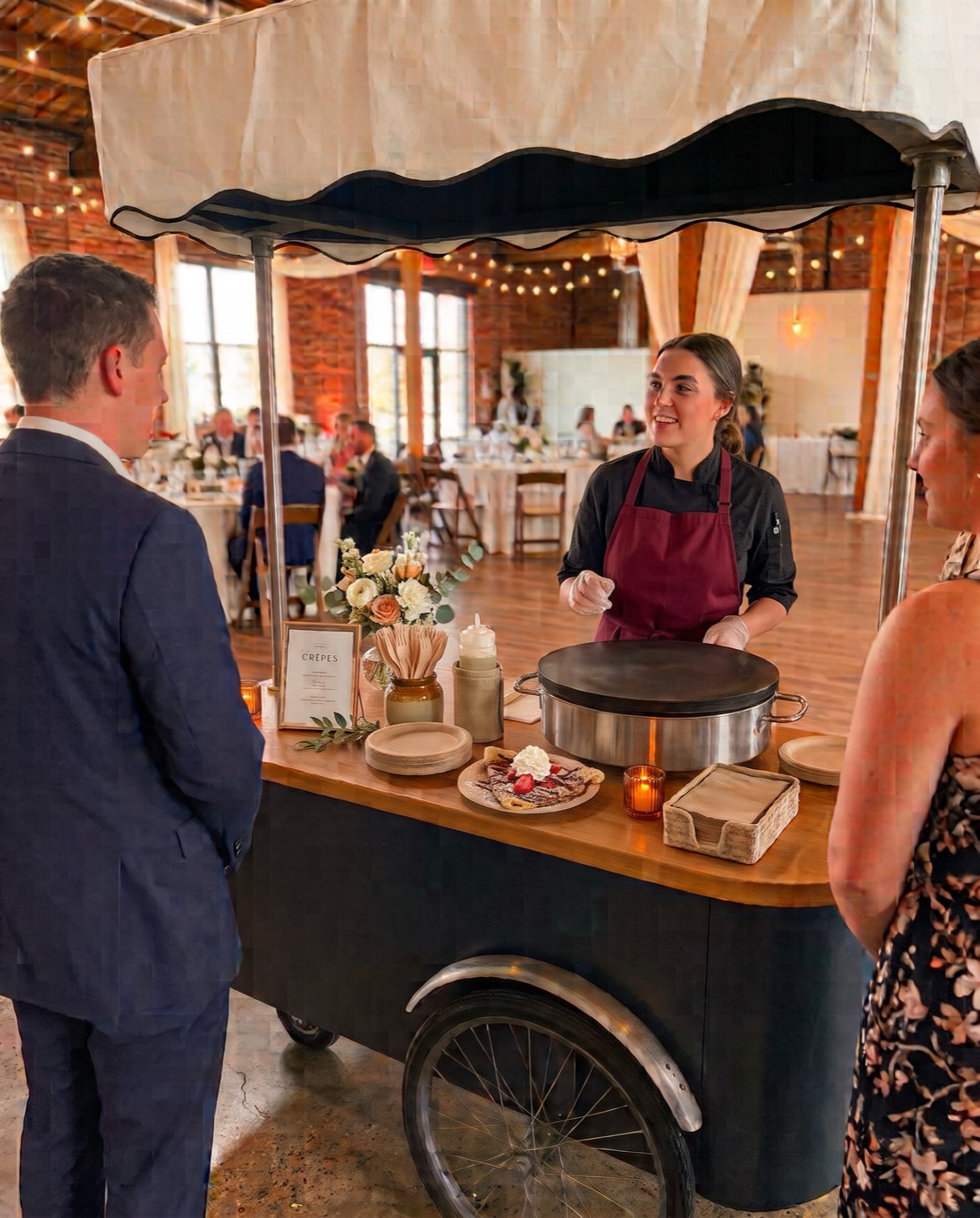 A Poppy Crêpes chef in a burgundy apron folding a crêpe at the cart inside a brick-walled reception venue, two guests waiting at the counter